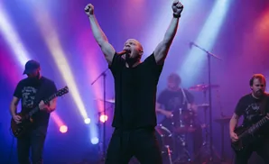 A band performs live on stage, bathed in purple and white spotlights. The lead singer is in the center with arms raised.