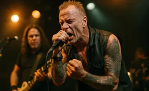 A male singer with tattoos in a black vest belts into a microphone on stage, with a guitarist behind him.