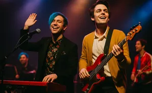 A band performs on stage, with two men in the foreground smiling and playing instruments.