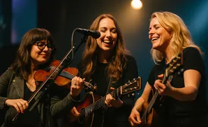 Three women playing music on stage: one plays the violin, and two play guitars.