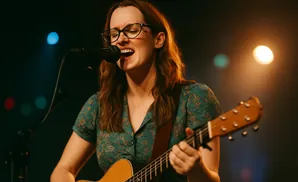 A woman with glasses sings into a microphone while playing an acoustic guitar.
