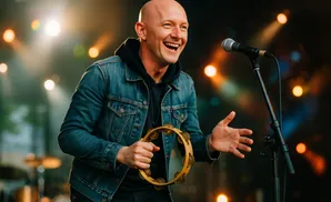 A bald man in a denim jacket laughs while holding a tambourine and singing into a microphone on stage.