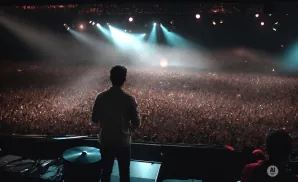 A DJ stands on stage facing a massive, cheering crowd with their hands in the air.