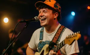 A smiling man in overalls and an orange baseball cap plays a guitar and sings into a microphone on stage.