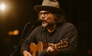 A man wearing a hat and glasses plays an acoustic guitar and sings into a microphone.
