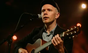 A man wearing a cap and suit plays an acoustic guitar and sings into a microphone.