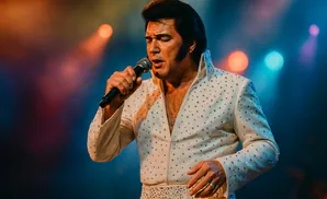An Elvis Presley impersonator in a white, rhinestone-covered jumpsuit sings into a microphone on a dimly lit stage with colorful lights in the background.
