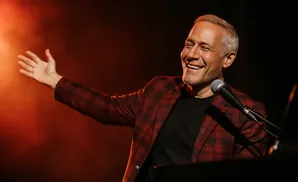 A man in a red plaid blazer smiles widely while reaching his hand out towards the left, with a microphone in front of him.