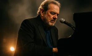 A man with gray hair and a beard plays the piano into a microphone.