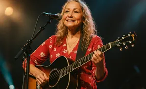 A woman with blonde hair smiles while playing an acoustic guitar and singing into a microphone on stage.