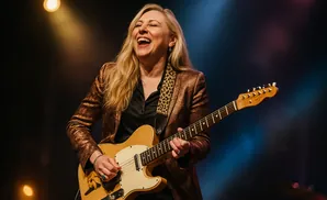 A woman with blonde hair laughs while playing a tan electric guitar on stage.