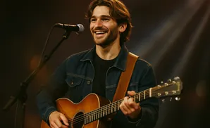 A smiling man with brown hair plays an acoustic guitar and sings into a microphone.