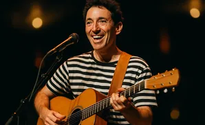 A man with a wide smile plays an acoustic guitar on stage, wearing a black and white striped shirt.