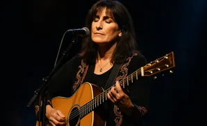 A woman with her eyes closed plays an acoustic guitar while singing into a microphone.