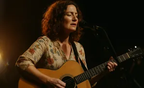 A woman with curly brown hair sings into a microphone while playing an acoustic guitar on a dimly lit stage.