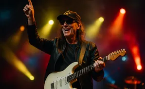 A man in a black leather jacket and cap plays a white electric guitar on stage, with colorful lights behind him.