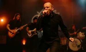 A bald man in a black shirt sings into a microphone on stage, with bandmates playing guitar and drums behind him.