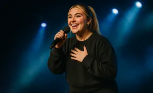 A woman with blonde hair in a ponytail sings into a microphone, holding her other hand to her chest.