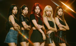 Five female K-pop idols in black outfits with microphones stand in a line, posing with their arms crossed.