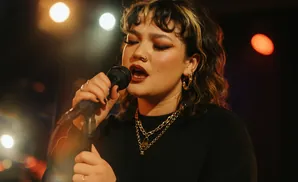 A woman with dark curly hair and gold hoop earrings sings into a microphone, wearing a black top and necklaces.
