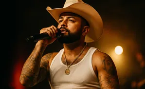 A man wearing a cowboy hat and white tank top sings into a microphone, with a gold chain around his neck.