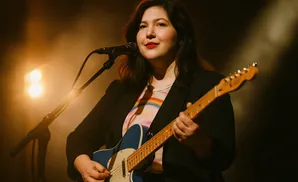 Woman playing a blue electric guitar on stage, lit by warm spotlights.