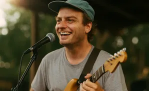 A man wearing a blue baseball cap smiles while singing into a microphone and playing an electric guitar.