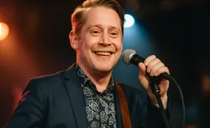 Macaulay Culkin smiles while holding a microphone and wearing a suit and floral shirt.