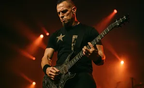 A male guitarist playing a black electric guitar on stage with orange spotlights.