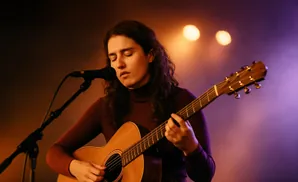Woman with long, curly hair plays an acoustic guitar and sings into a microphone under stage lights.