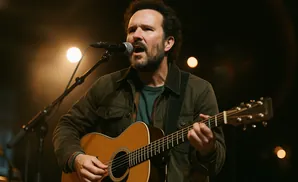 A man sings and plays an acoustic guitar on stage with warm lighting.