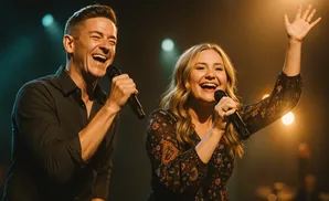 A man and woman singing joyfully into microphones on a stage with warm lighting.