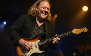 A man with long blonde hair laughs as he plays a sunburst Fender Stratocaster guitar on stage.