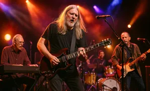 Guitarist with long blonde hair sings while playing a Gibson SG guitar on stage.