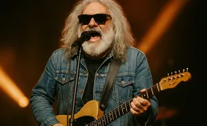 A man with long gray hair, sunglasses, and a beard sings into a microphone while playing a guitar.
