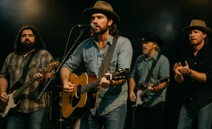 A band performs on stage with the lead singer in the foreground playing an acoustic guitar and singing into a microphone.