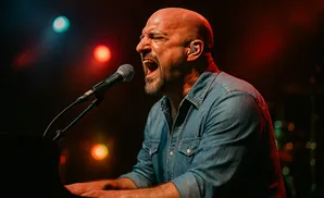 A bald man with a beard sings into a microphone while playing a piano on a dimly lit stage.