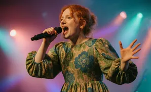 Singer with red hair, wearing a floral dress, singing into a microphone on a stage with colored lights.