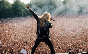 Singer with long blonde hair in a black studded jacket and pants, singing into a microphone with a large crowd cheering.