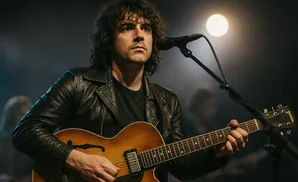 A man with curly hair plays an acoustic guitar on stage, wearing a black leather jacket.
