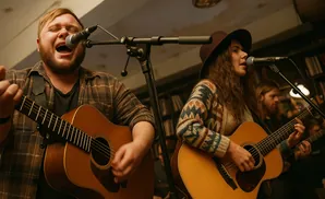 Two musicians, a man and a woman, play acoustic guitars and sing into microphones.