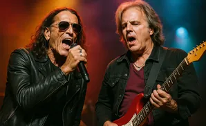 Two male rock musicians perform on stage, one singing into a microphone, the other playing a red electric guitar.
