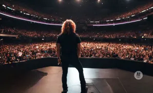 A musician with long, curly red hair stands on stage facing a massive, cheering crowd in a stadium.