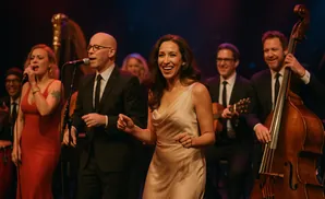 A diverse band performs on stage with a female lead singer in a cream dress dancing.