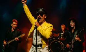 A Freddie Mercury impersonator in a yellow jacket sings into a microphone with a band behind him.