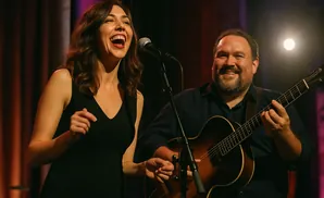 A woman singing into a microphone while a man plays guitar next to her.