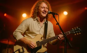 A blond, curly-haired musician smiles broadly while playing a cream-colored electric guitar under warm stage lights.