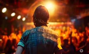 A person in a plaid shirt faces a bright stage light in a blurry, energetic crowd.