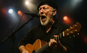 Musician in a black beret and shirt sings into a microphone while playing an acoustic guitar on stage.