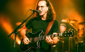 A man with long hair and glasses plays a black electric guitar and sings into a microphone on stage.